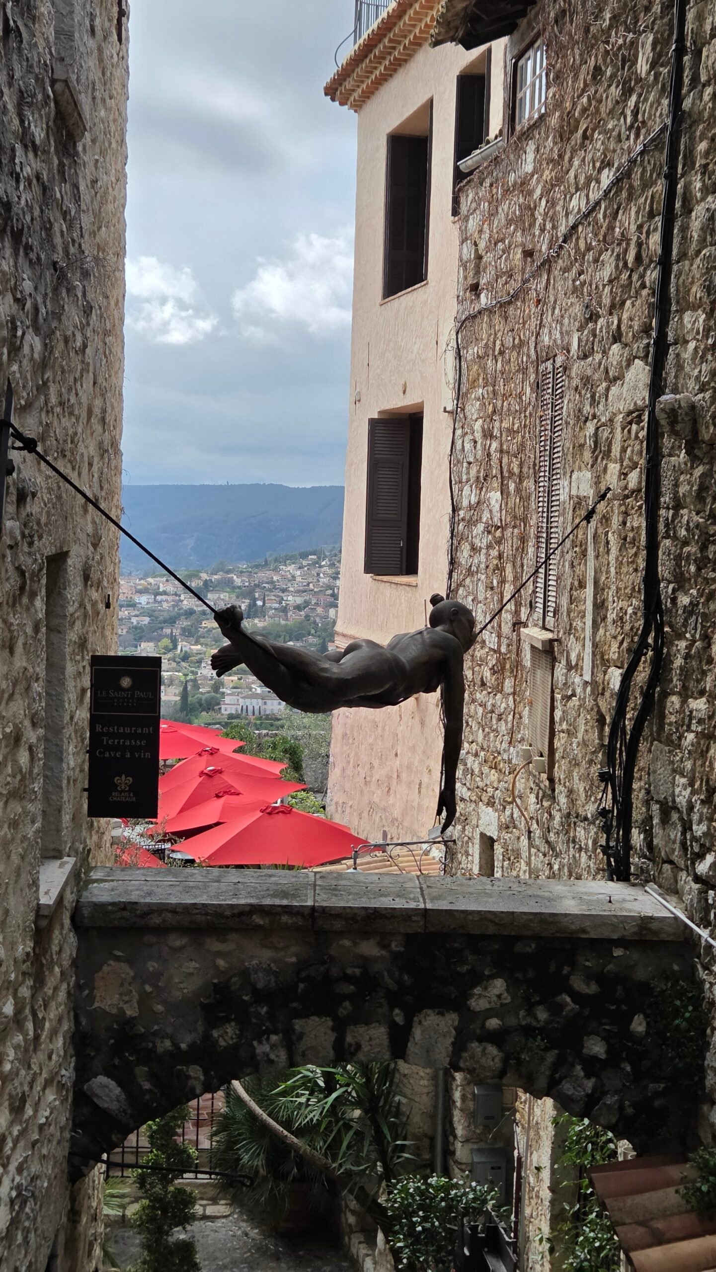 Bronze sculpture of a flying figure suspended between buildings over an archway in Saint-Paul-de-Vence